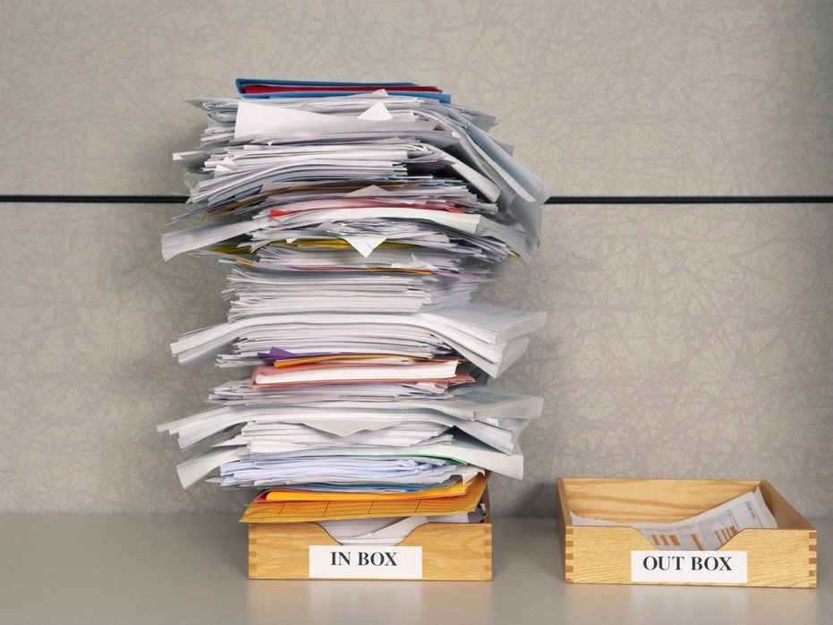 A tall stack of papers on a desk next to wooden trays labelled in box and out box