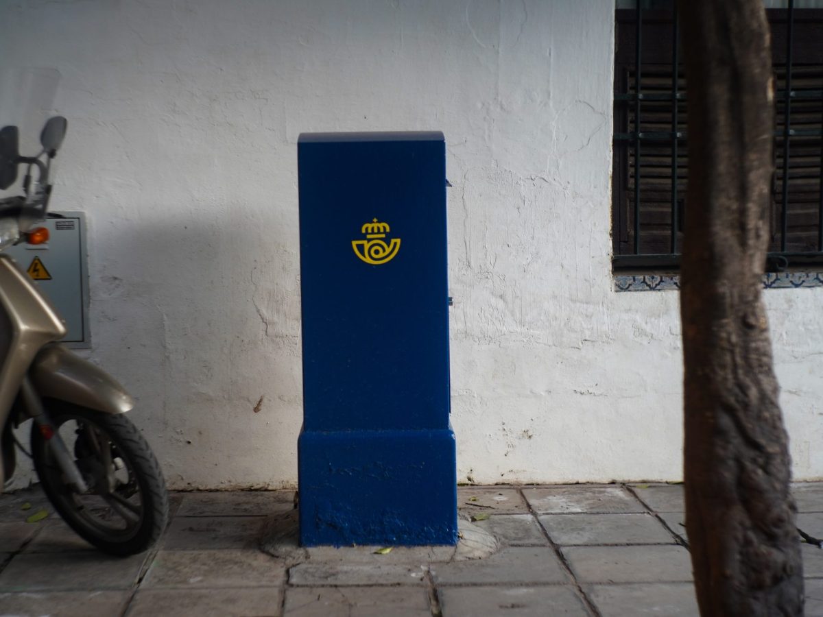 A square deep blue mailbox on a street under a tree. Ein quadratischer dunkelblauer Briefkasten an einer Straße unter einem Baum.